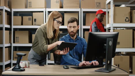 Man-And-Woman-Working-Together-At-Computer-While-Entering-Data-From-Tablet-Device-In-Postal-Office