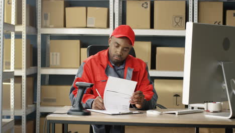 Young-Handsome-Postman-In-Uniform-And-Cap-Sitting-At-Computer-And-Filling-In-Invoice-Of-Parcel