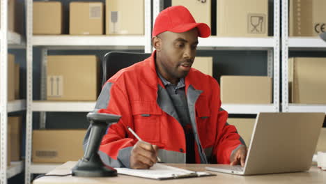 Junger-Postbote-In-Roter-Uniform-Und-Mütze,-Der-Am-Schreibtisch-Im-Postgeschäft-Sitzt-Und-Am-Laptop-Arbeitet