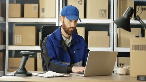 Schöner-Postbote-In-Uniform-Und-Mütze,-Der-Am-Schreibtisch-Im-Postgeschäft-Sitzt-Und-Am-Laptop-Arbeitet