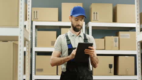 Handsome-Young-Postman-Standing-In-Postal-Store-With-Parcels-And-Tapping-On-Tablet-Computer