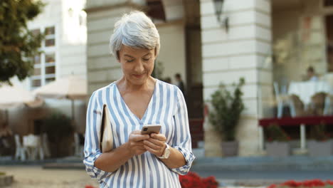 Beautiful-Old-Woman-Standing-In-Center-City-And-Tapping-Message-On-Smartphone