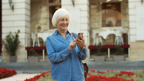 Schöne-Alte-Frau,-Die-In-Der-Stadtmitte-Steht-Und-Auf-Dem-Smartphone-Auf-Eine-Nachricht-Tippt-1