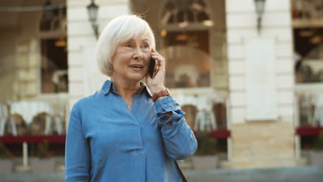 Grey-Haired-Old-Woman-Standing-In-City-And-Chatting-On-Smartphone