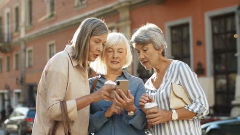 Three-Beautiful-And-Cheerful-Women-With-Coffee-To-Go-In-Hands-Laughing-While-Scrolling-Photos-On-Smartphone-Outdoor