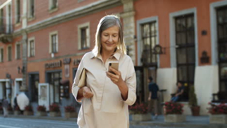 Senior-Good-Looking-Lady-Walking-The-Street-In-Old-City-And-Tapping-On-Smartphone