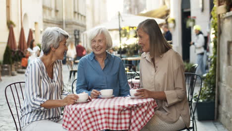 Three-Senior-Women-Friendly-Chatting-And-Drinking-Coffee-At-Table-In-Cafe-Terrace-In-Summer