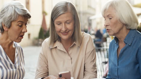 Drei-ältere-Frauen,-Die-Freundlich-Auf-Das-Smartphone-Schauen-Und-Im-Sommer-Kaffee-Am-Tisch-Auf-Der-Caféterrasse-Trinken