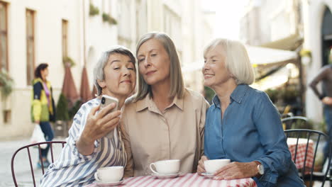 Drei-Hübsche-ältere-Freundinnen-Sitzen-Mit-Kaffee-Am-Tisch-Auf-Der-Caféterrasse-Und-Lächeln-In-Die-Smartphone-Kamera,-Während-Sie-Selfie-Fotos-Machen
