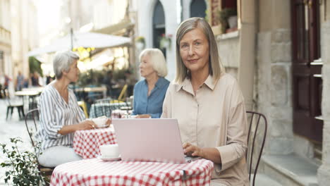 Portrait-Of-Beautiful-Old-Woman-With-Gray-Hair-Typing-On-Laptop-Computer-And-Smiling-To-Camera-At-Table-In-Cafe-Outdoors