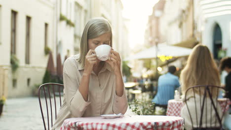 Good-Looking-Senior-Smiled-Woman-With-Gray-Hair-Sipping-Coffee-While-Sitting-At-Table-In-Cafe-Terrace-Outdoors
