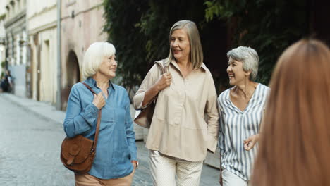 Three-Cheerful-Senior-Women-On-Retirement-Walking-The-Old-City-Street-And-Laughing-While-Talking