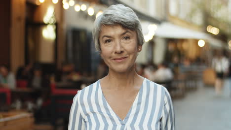 Portrait-Of-Beautiful-Senior-Woman-With-Grey-Hair-Looking-Straight-At-Camera-And-Smiling-Cheerfully-Outdoor-In-Town