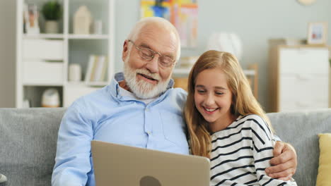 Portrait-Shot-Of-The-Pretty-Teenage-Girl-Laughing-Together-With-Her-Grandfather-While-They-Looking-At-The-Screen-Of-Laptop-Computer-In-The-Nice-Living-Room