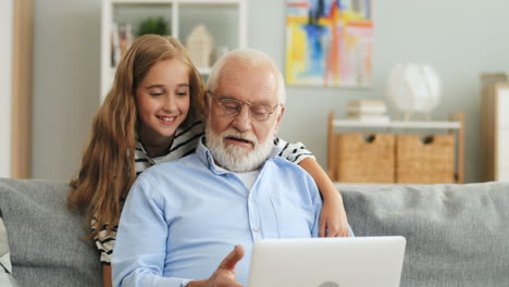Pretty-Teen-Girl-Hugging-Her-Grandfather-From-Behind-While-He-Sitting-On-The-Sofa-And-Typing-On-The-Laptop-At-Home