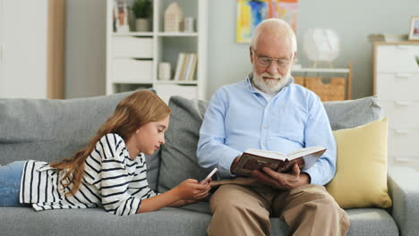 Der-Grauhaarige-Alte-Großvater-In-Der-Brille-Liest-Ein-Buch-Und-Seine-Enkelin-Benutzt-Das-Smartphone,-Während-Sie-Sich-Auf-Der-Couch-Ausruhen