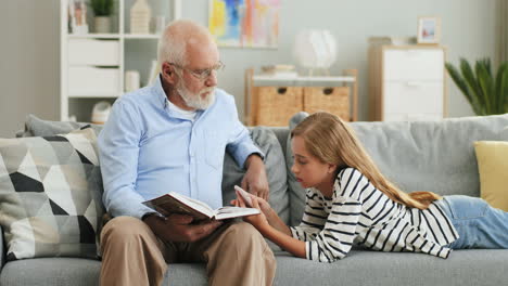 Senior-Smile-Grandpa-Reading-A-Book-For-His-Granddaughter-While-She-Holding-A-Smartphone-On-The-Sofa-In-The-Living-Room