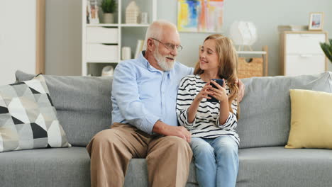 Grandfather-Embracing-His-Pretty-Granddaughter-While-They-Sitting-On-The-Sofa-In-The-Living-Room-And-Girl-Tapping-On-The-Smartphone-Screen-And-Man-Watching-At-It