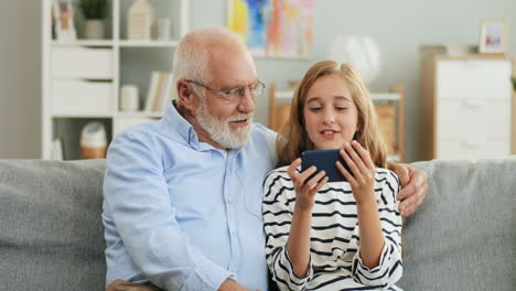 Portrait-Shot-Of-The-Grandfather-And-Granddaughter-Resting-On-The-Couch-In-The-Living-Room-And-Girl-Showing-Something-To-The-Grandpa-On-The-Phone-Screen