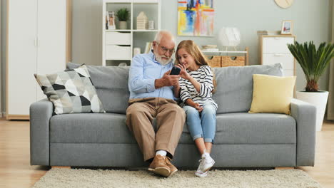 älterer-Grauhaariger-Opa-Mit-Brille,-Der-Seiner-Enkelin-Etwas-Auf-Dem-Telefonbildschirm-Demonstriert,-Während-Sie-Im-Gemütlichen-Wohnzimmer-Sitzen