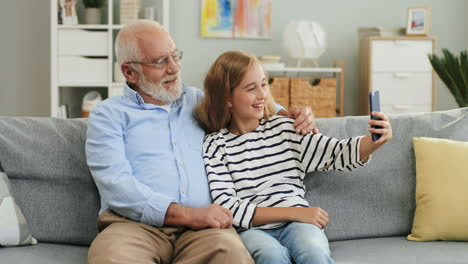 Portrait-Shot-Of-The-Happy-Smiled-Grandfather-And-Teenage-Granddaughter-Taking-Selfie-Photos-While-Sitting-On-The-Couch-At-Home
