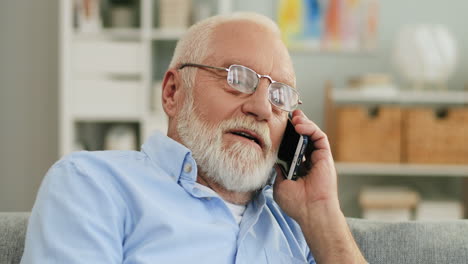 Close-Up-Of-The-Senior-Man-With-Grey-Hair-And-In-Glasses-Talking-On-The-Phone-And-Smiling-While-Sitting-In-The-Cozy-Living-Room