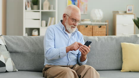 Old-Grey-Haired-Man-In-Glasses-Sitting-On-The-Couch-At-Home-And-Texting-A-Message-On-The-Smartphone
