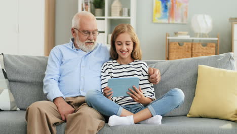 Grandfather-Embracing-His-Pretty-Granddaughter-While-They-Sitting-On-The-Sofa-In-The-Living-Room-And-Girl-Tapping-On-The-Tablet-Device-Screen-And-Man-Watching-At-It