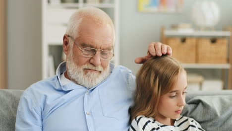 Close-Up-Of-The-Old-Grandpa-Reading-A-Book-And-Caressing-His-Granddaughter-On-The-Head-While-She-Sitting-Beside-With-A-Smartphone