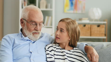Portrait-Of-The-Smiled-Grandpa-In-Glasses-And-Blonde-Pretty-Grandaughter-Sitting-Close-Together-On-The-Sofa-In-The-Cozy-Room