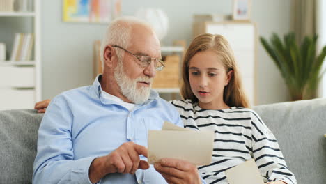 Portrait-Shot-Of-The-Old-Grey-Haired-Man-In-Glasses-Showing-Old-Photographs-To-His-Granddaughter-While-They-Sitting-On-The-Sofa