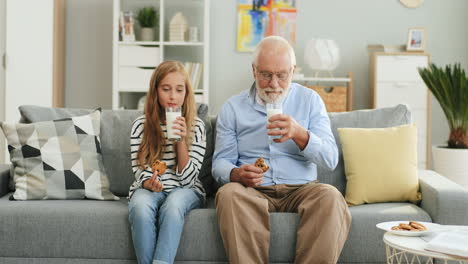 Pretty-Blonde-Teen-Girl-Sitting-Together-With-Her-Grandfather-With-Grey-Haired-On-The-Sofa-At-Home-And-Drinking-Milk-With-Cookies