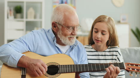 Portrait-Shot-Of-The-Old-Grandfather-In-Glasses-Playing-The-Guitar-And-His-Granddaughter-Listening-To-Him-At-Home