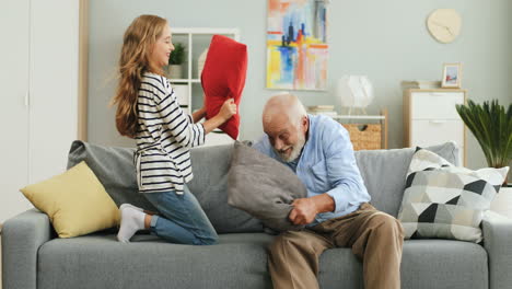 Cute-Teenage-Girl-Having-A-Pillows-Fight-With-Her-Old-Grandfather-On-The-Sofa-In-The-Cozy-Room