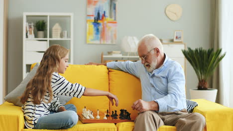 Senior-Man-With-Grey-Hair-Playing-Chess-With-His-Cute-Teen-Granddaughter-On-The-Sofa-At-Home