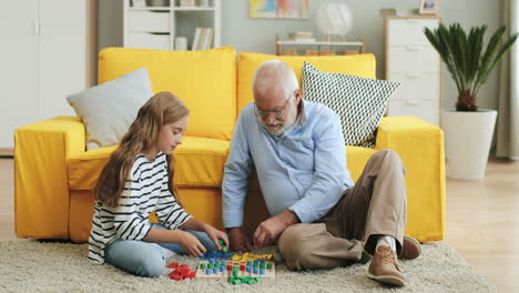 Senior-Grandfather-With-Grey-Hair-Playing-A-Game-With-His-Cute-Teen-Granddaughter-On-The-Sofa-At-Home