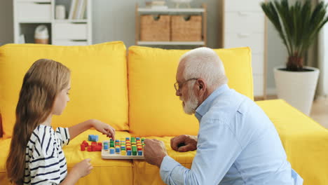 Close-Up-Of-The-Old-Grandpa-In-Glasses-Playing-A-Game-With-His-Pretty-Teen-Granddaughter-On-The-Yellow-Couch-At-Home