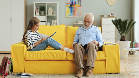Pretty-Teen-Schoolgirl-Doing-Her-Homework-Together-With-Her-Grandfather-In-Glasses-On-The-Yellow-Sofa-In-The-Living-Room