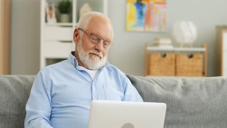 älterer-Mann-Mit-Grauen-Haaren-Und-Brille-Sitzt-Auf-Der-Couch,-Sieht-Sich-Etwas-Auf-Dem-Laptop-An-Und-Lächelt