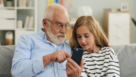 Portrait-Of-The-Cute-Blond-Teen-Girl-And-His-Grey-Haired-Grandpa-Who-Showing-Her-Something-On-The-Smartphone-Screen-Sitting-On-The-Couch-At-Home