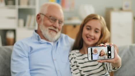 Portrait-Of-The-Pretty-Schoolgirl-Sitting-On-The-Sofa-With-A-Grandfather-In-Glasses,-Smiling-And-Taking-Selfie-Photo-On-The-Smartphone-Camera