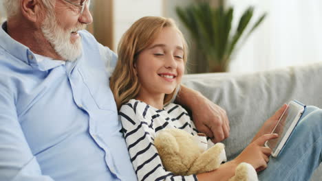 Close-Up-Of-The-Senior-Grandfather-In-Glasses-Sitting-On-The-Couch-And-Hugging-His-Pretty-Teen-Granddaughter-Who-Holding-Teddy-Bear-And-Scrolling-On-The-Tablet-Device