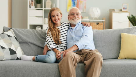Portrait-Shot-Of-The-Smiled-Happy-Granddaughter-And-Grandfather-Hugging-And-Posing-To-The-Camera-While-Sitting-On-The-Sofa-In-The-Cozy-Room