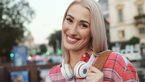 Close-Up-Of-The-Attractive-Joyful-Young-Blond-Woman-Tourist-With-Headphones-And-Backpack-Smiling-While-Standing-In-The-City-Center