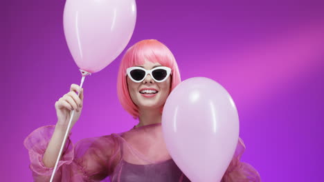 Portrait-Shot-Of-Happy-Stylish-Young-Attractive-Woman-In-Pink-Wig-And-Glasses-Dancing-With-Balloons-And-Smiling-To-Camera-On-Bright-Wall-Background