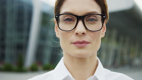 Close-Up-Of-The-Charming-Woman-In-Glasses-Looking-Straight-Into-The-Camera-On-The-Big-Modern-Glass-Building-Background
