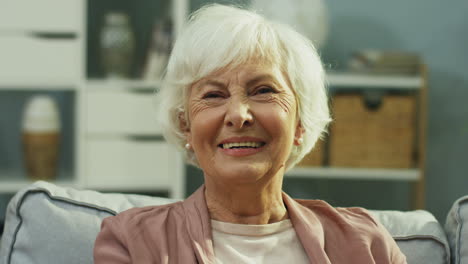 Close-Up-Of-The-Old-Lady-On-Retirement-Smiling-To-The-Camera-While-Sitting-On-The-Gray-Couch-In-The-Living-Room