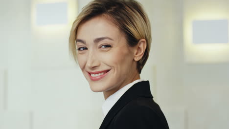 Side-View-Portrait-Of-The-Young-Attractive-Businesswoman-With-Short-Fair-Hair-Turning-Her-Head-To-The-Camera-And-Smiling-Cheerfully-To-The-Camera-On-The-Wall-Background