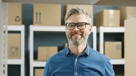 Close-Up-Of-Handsome-Postman-In-Glasses-Taking-Carton-Box-With-Bar-Code-From-Shelf-And-Smiling-In-Post-Office-Store