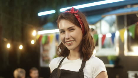 Close-Up-Of-Beautiful-Young-Woman-In-Apron-Smiling-Happily-To-Camera-Outdoors-With-Little-Food-Truck-Bar-On-Background